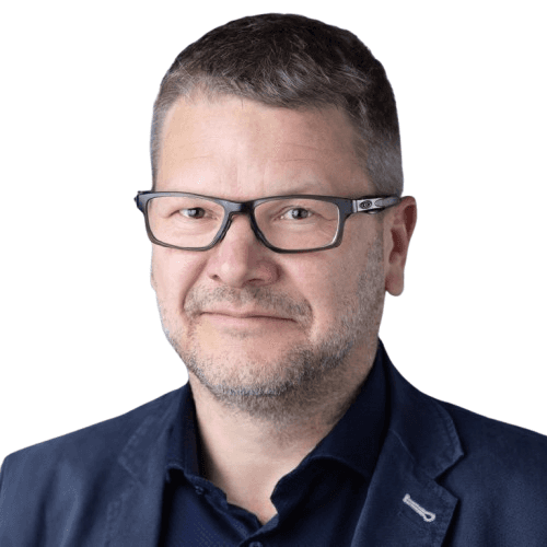 A smiling man with curly hair is sitting at a desk, looking at the camera, with a laptop and a lamp in the background.