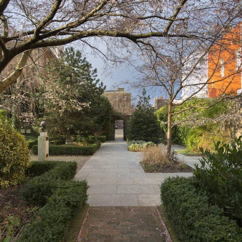 A garden pathway lined with hedges and trees, leading to a building in the background. Bust sculpture is visible on the left.