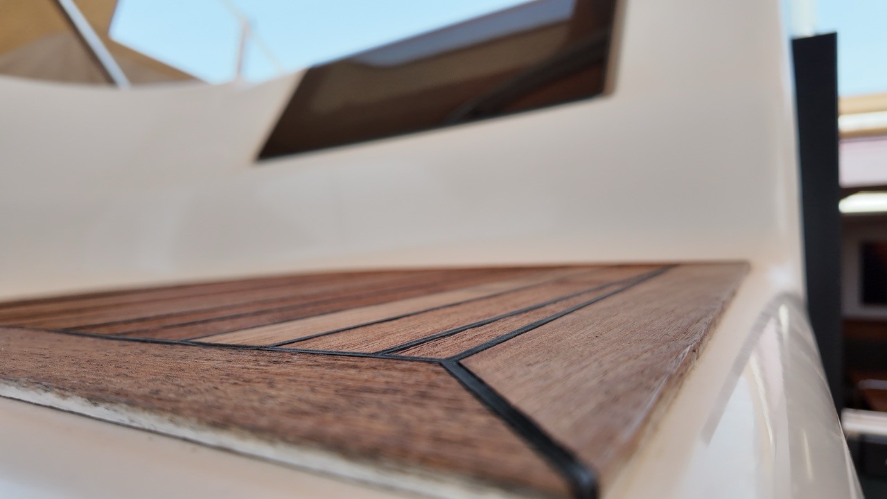 Close-up of the Hanse 575's foredeck showing a herringbone teak inlay panel set into the white deck, beside a hatch and stainless steel rail fitting.