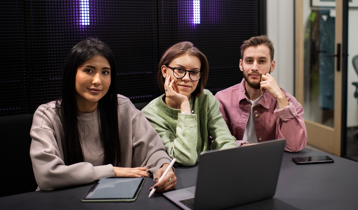 Three people are seated at a conference table with a laptop and tablet, engaging in a casual meeting or discussion in a modern office setting.