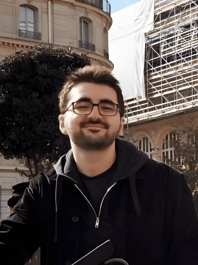 A person stands outdoors in a natural setting with lush green trees under a clear sky, wearing a dark sweatshirt, and looking into the distance.