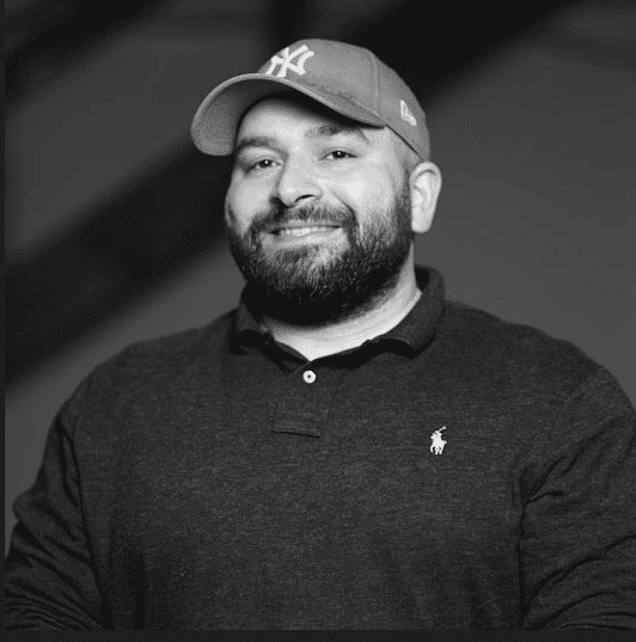 A smiling man with a beard and a baseball cap stands against a dark background, wearing a black polo shirt.