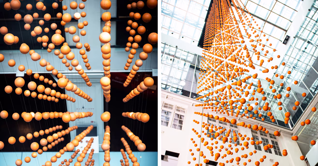 A split image of a kinetic sculpture featuring numerous suspended orange spheres. The left side shows a bottom-up view, while the right side displays a wider perspective of the installation against a glass-ceilinged interior.