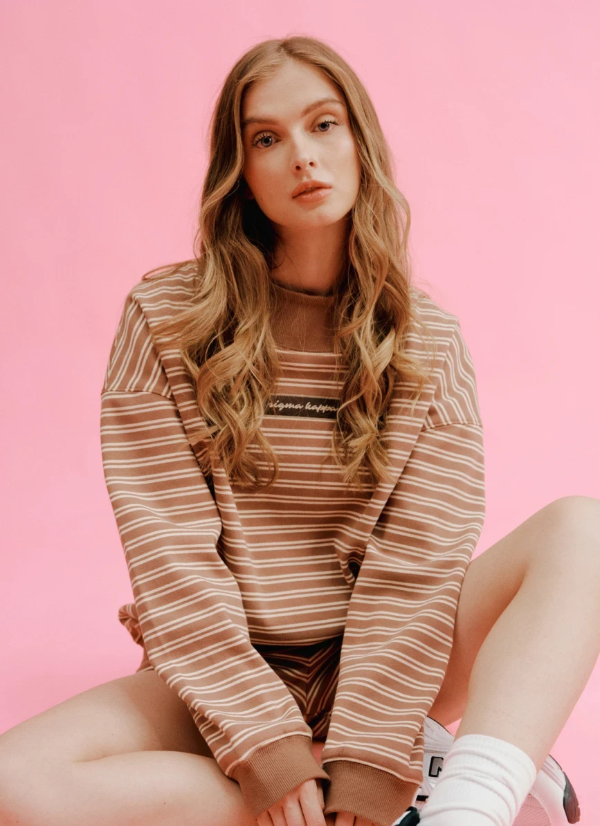 Model wearing an oversized brown-and-cream striped sorority sweatshirt against a pink background.