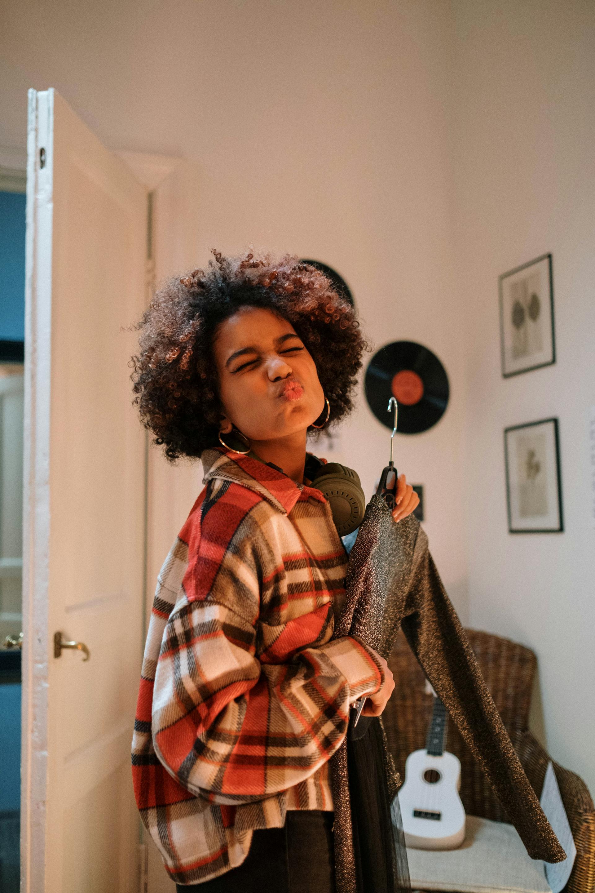 A young woman wearing a red plaid shirt and headphones making a "kiss" face while holding a sparkly garment on a hanger.