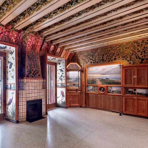 Bogato ukrašena prostorija s cvjetnim i mozaičnim zidnim uzorcima, drvenim ormarićima, kaminom i velikim prozorima, uz uokvirenu sliku pejzaža.