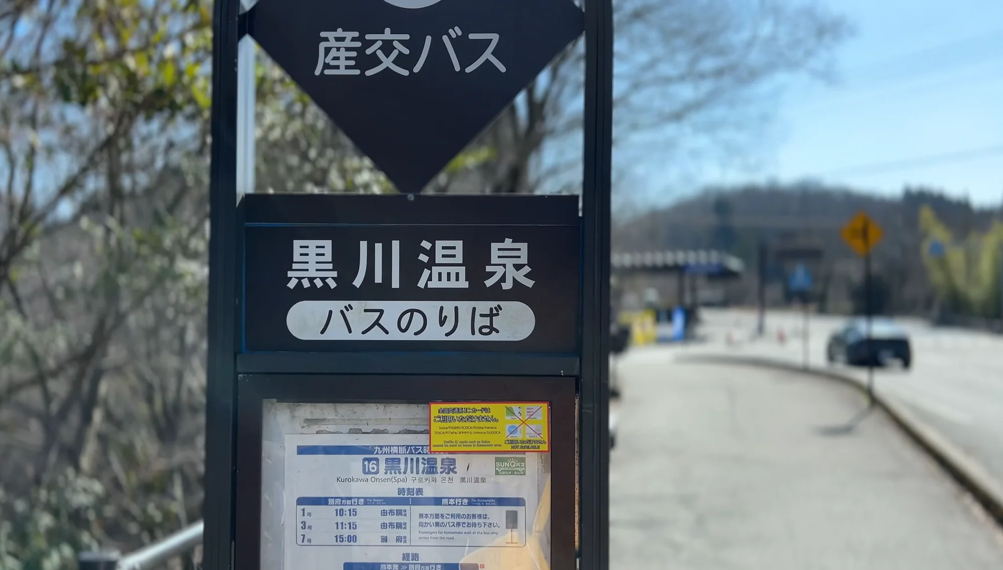 A Sanko bus stop sign reading Kurokawa Onsen with a timetable case at the roadside