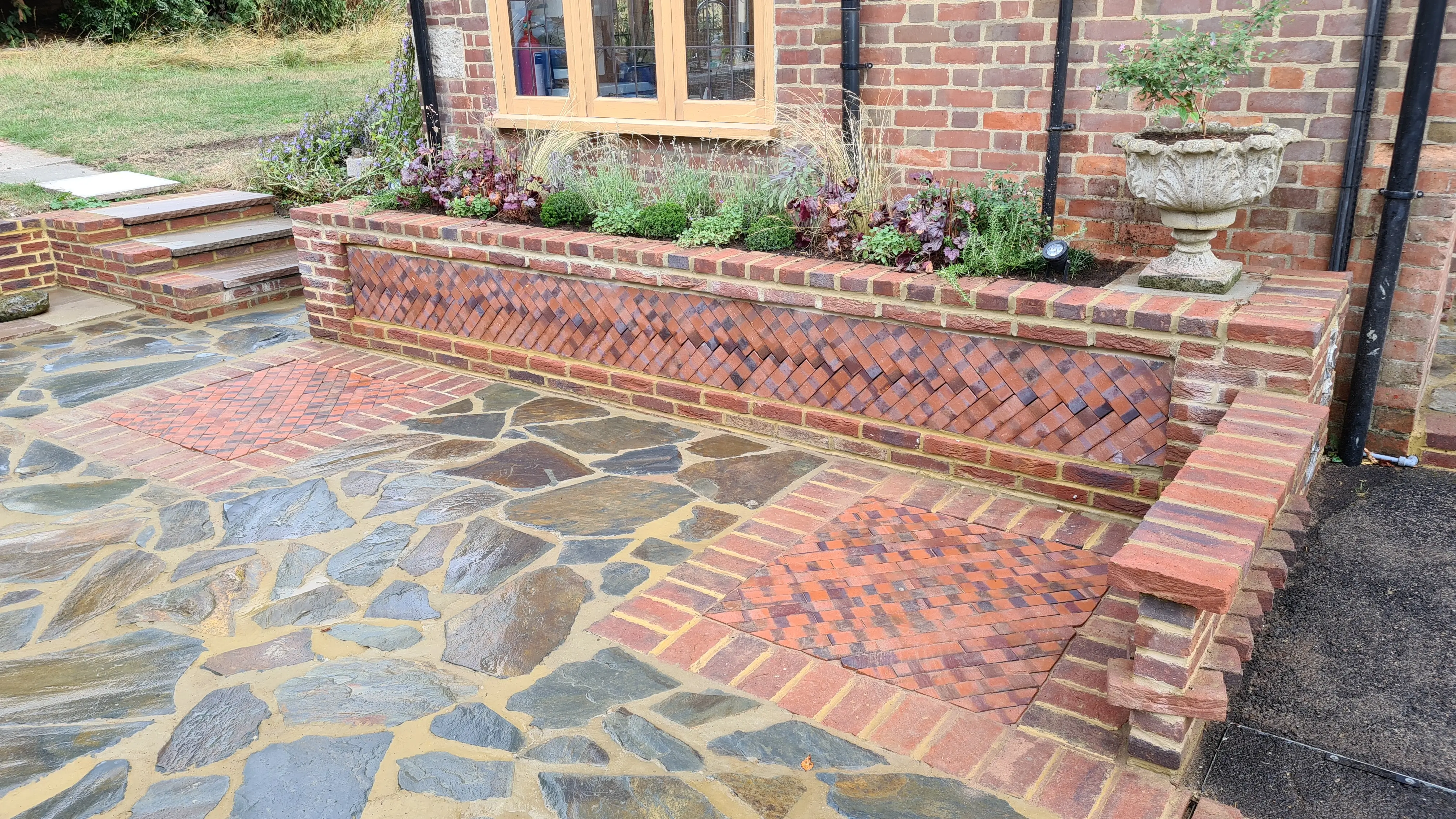 A stone patio featuring a brick seating area with plants in the background near a house.