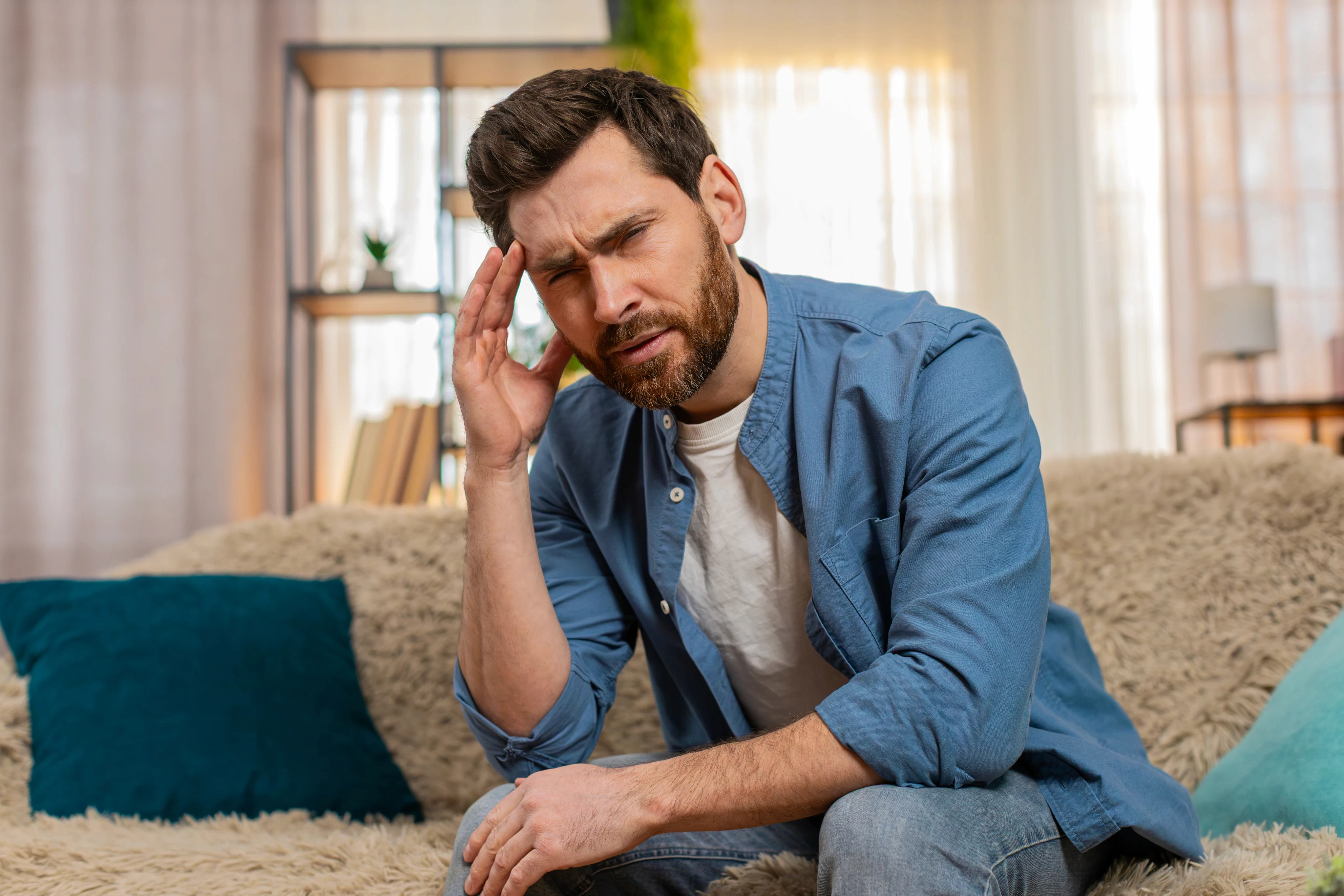Man in a blue shirt sits on a couch, holding his head with a pained expression. Cozy living room background with pillows and a bookshelf.