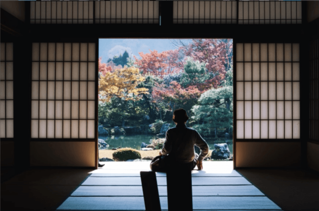 Silhouettes of a parson sitting on a wooden floor viewing a vibrant autumn garden through open shoji doors in a traditional Japanese room