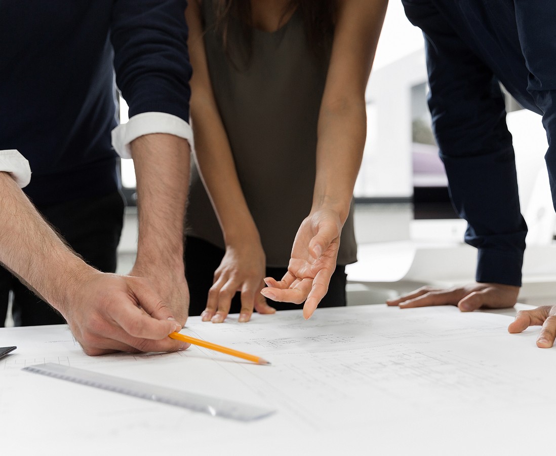 Three individuals collaborate over a large desk, discussing architectural blueprints, with one person pointing to a specific part of the plan using a pencil, surrounded by drafting tools including a ruler.
