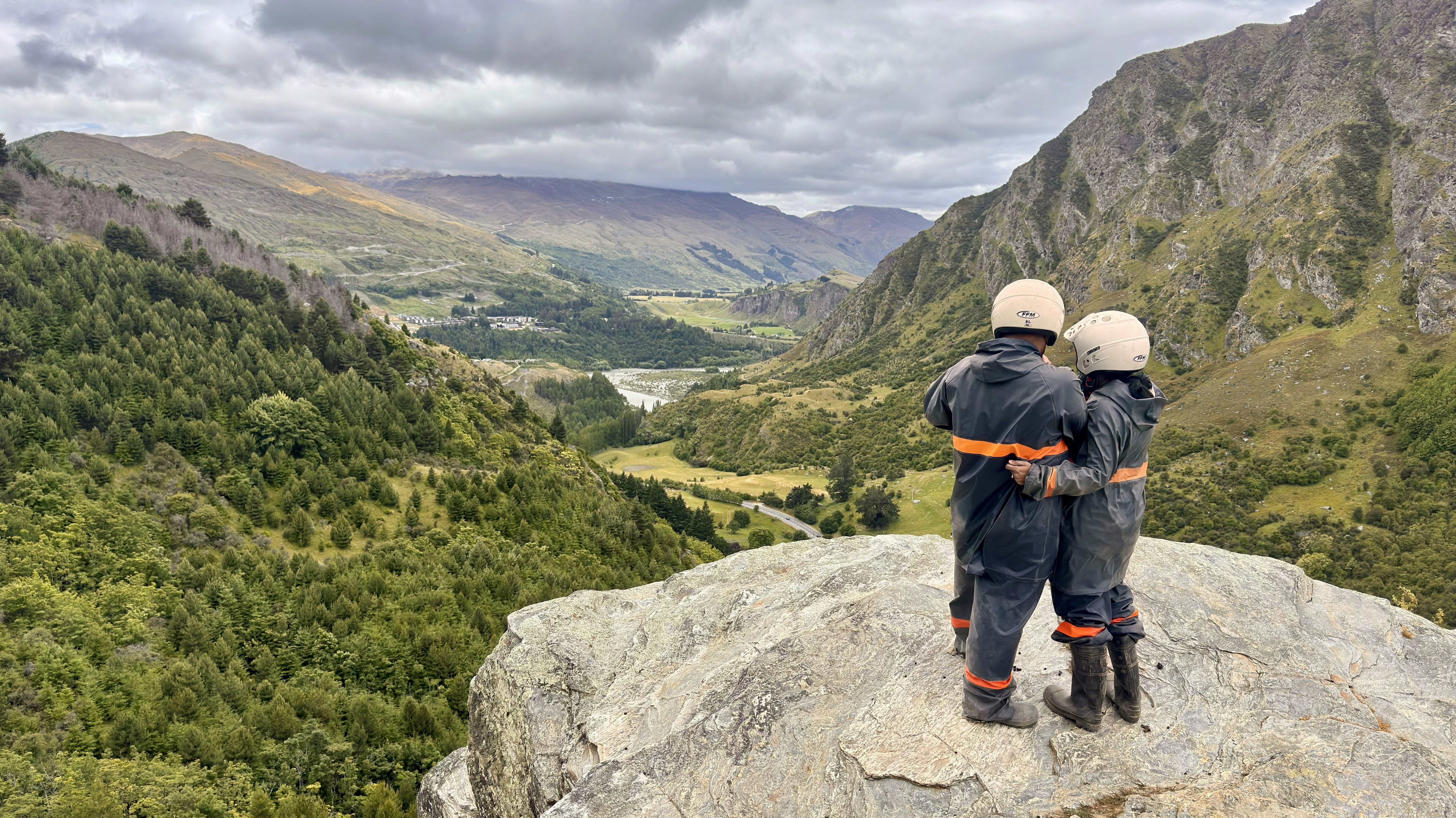 A couple wearing a gear and helmet, standing on the rock facing a beautiful valley in Queenstown