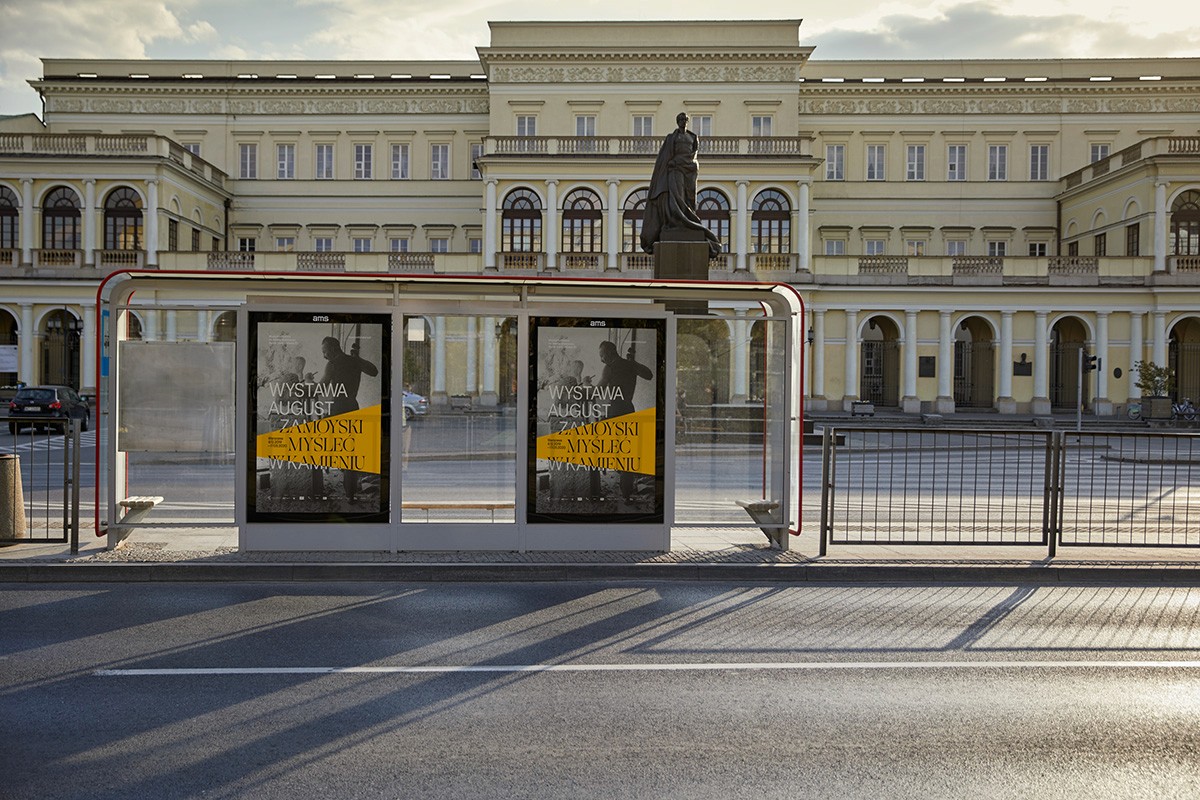 Two exhibition posters displayed in a minimalist glass bus shelter at sunset, featuring high-contrast yellow and black graphics. This illustrates a comprehensive out-of-home (OOH) marketing strategy for a major cultural event.