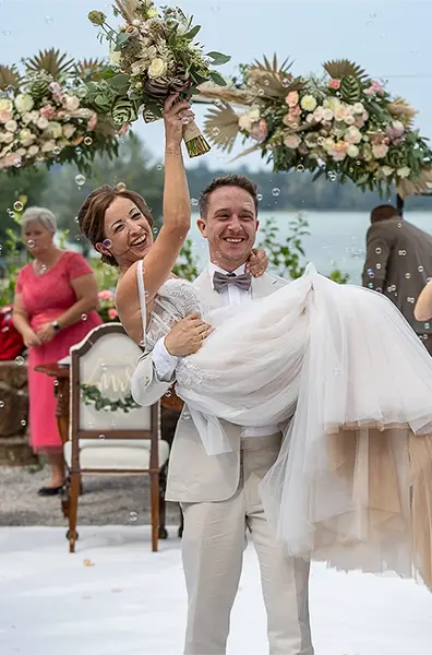 Lächelnder Bräutigam trägt Braut, Schloss Mattsee, mit Brautstrauß. Bubbles.