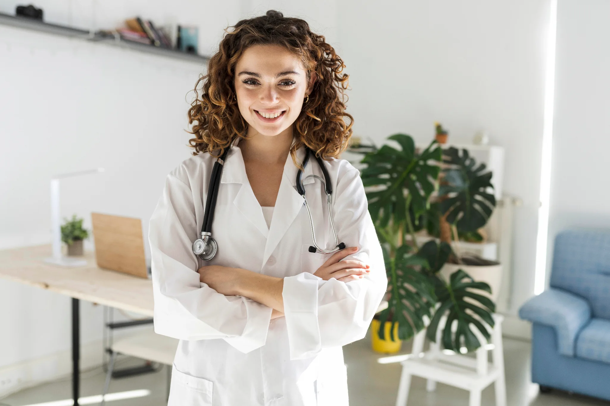 A smiling female doctor with curly auburn hair wearing a white lab coat and stethoscope stands with crossed arms in a bright, modern medical office decorated with potted plants.