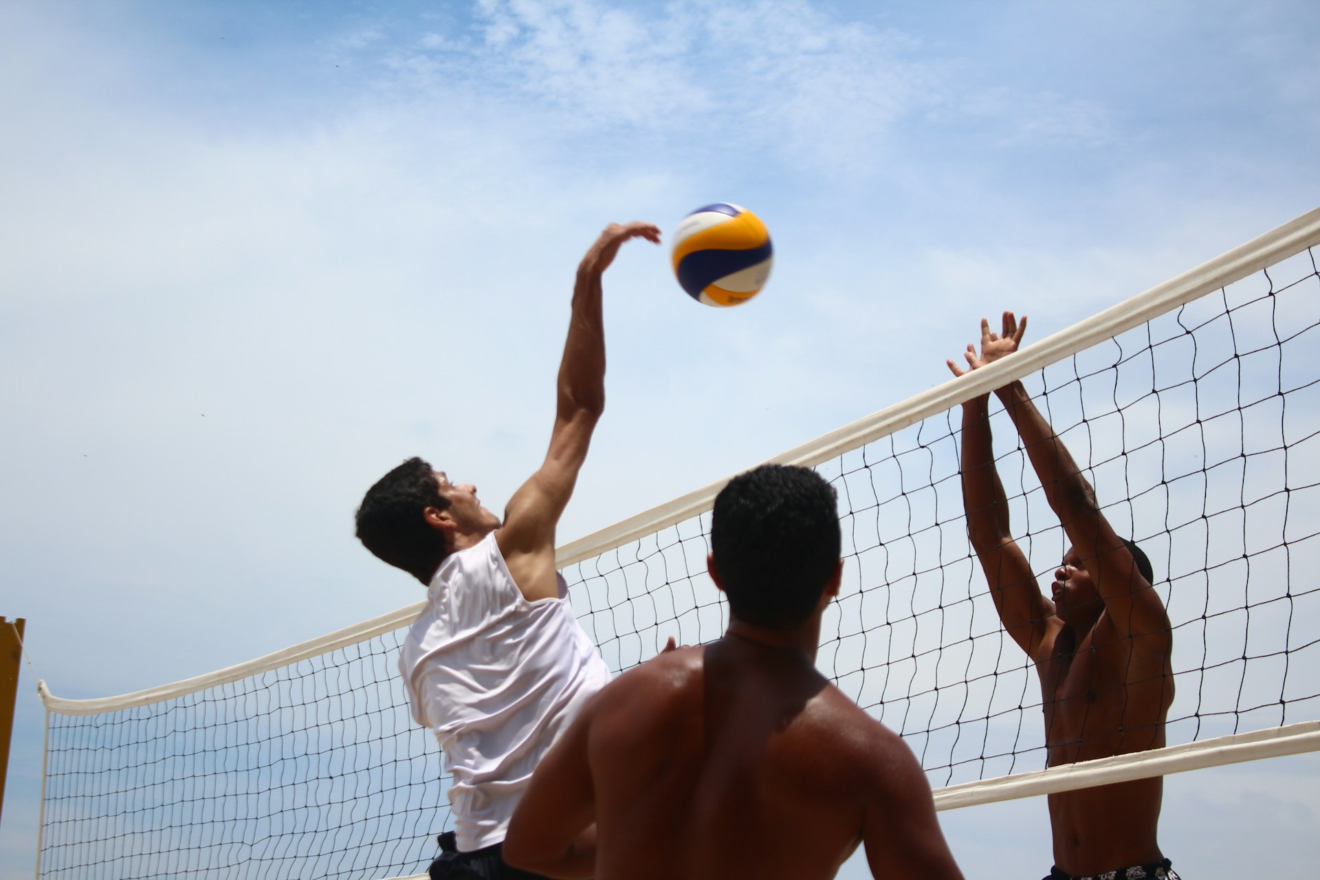 A beach volleyball player in a white tank top jumping to spike the ball against two opposing blockers.