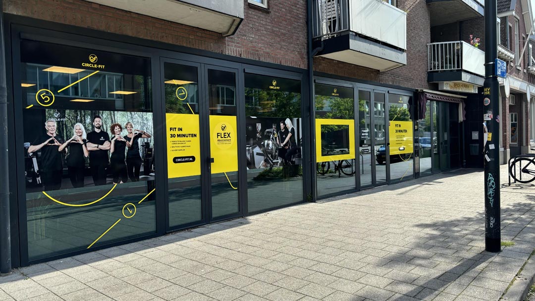 A Circle Fit hub with large windows displaying bright yellow signs and promotions, viewed from the sidewalk.