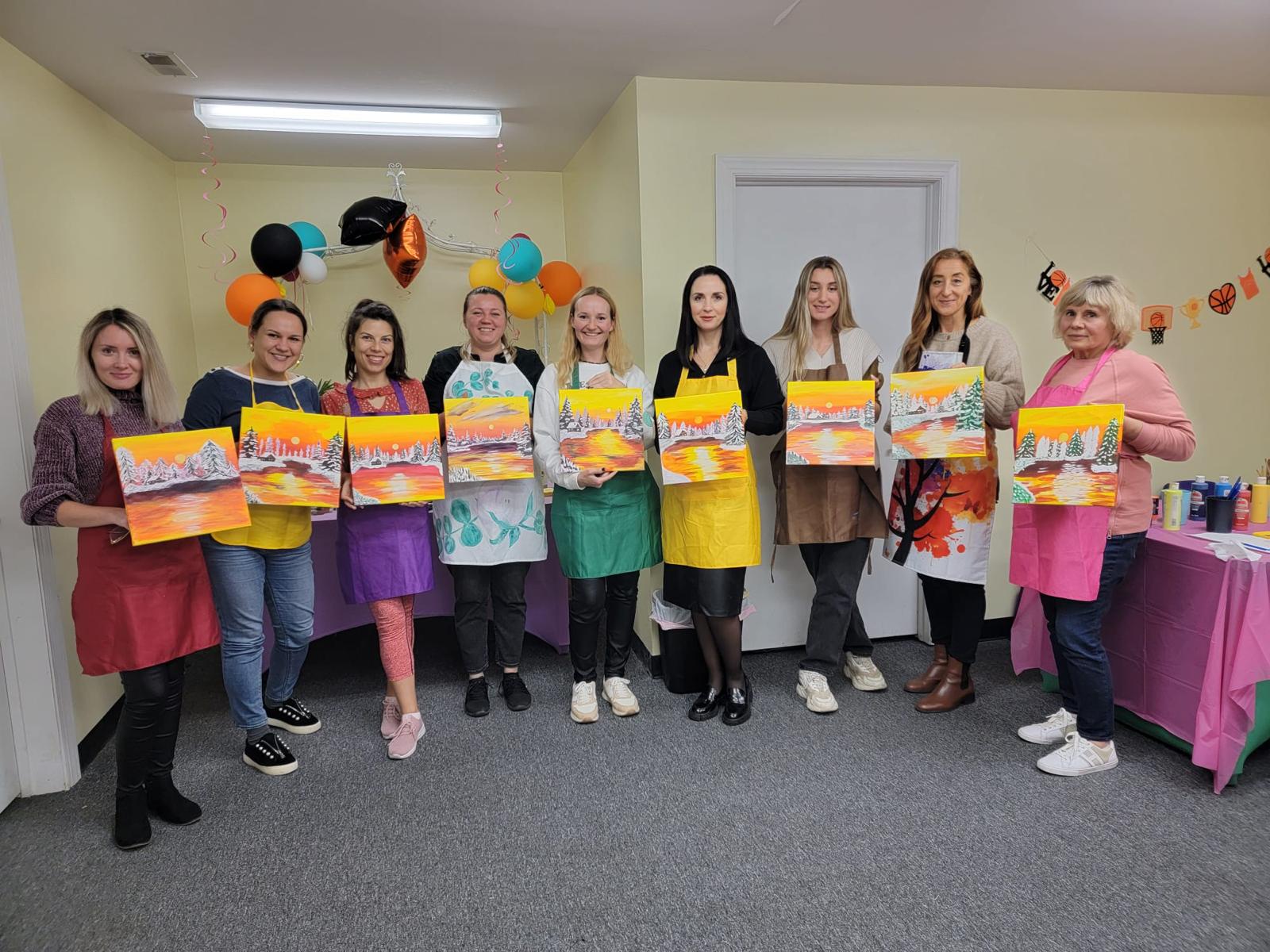 Nine women lined up showing their paintings from a recent SportPlus Paint & Sip Class