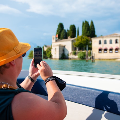 Person in a yellow hat taking a photo with a smartphone from a boat, capturing buildings and trees across a body of water.