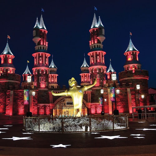 Isang iluminadong kastilyo na may mga tore sa gabi, na may gitnang gintong estatwa ng isang pigura na nakataas ang mga kamay sa harap. Puti ang mga pattern ng bituin sa lupa.