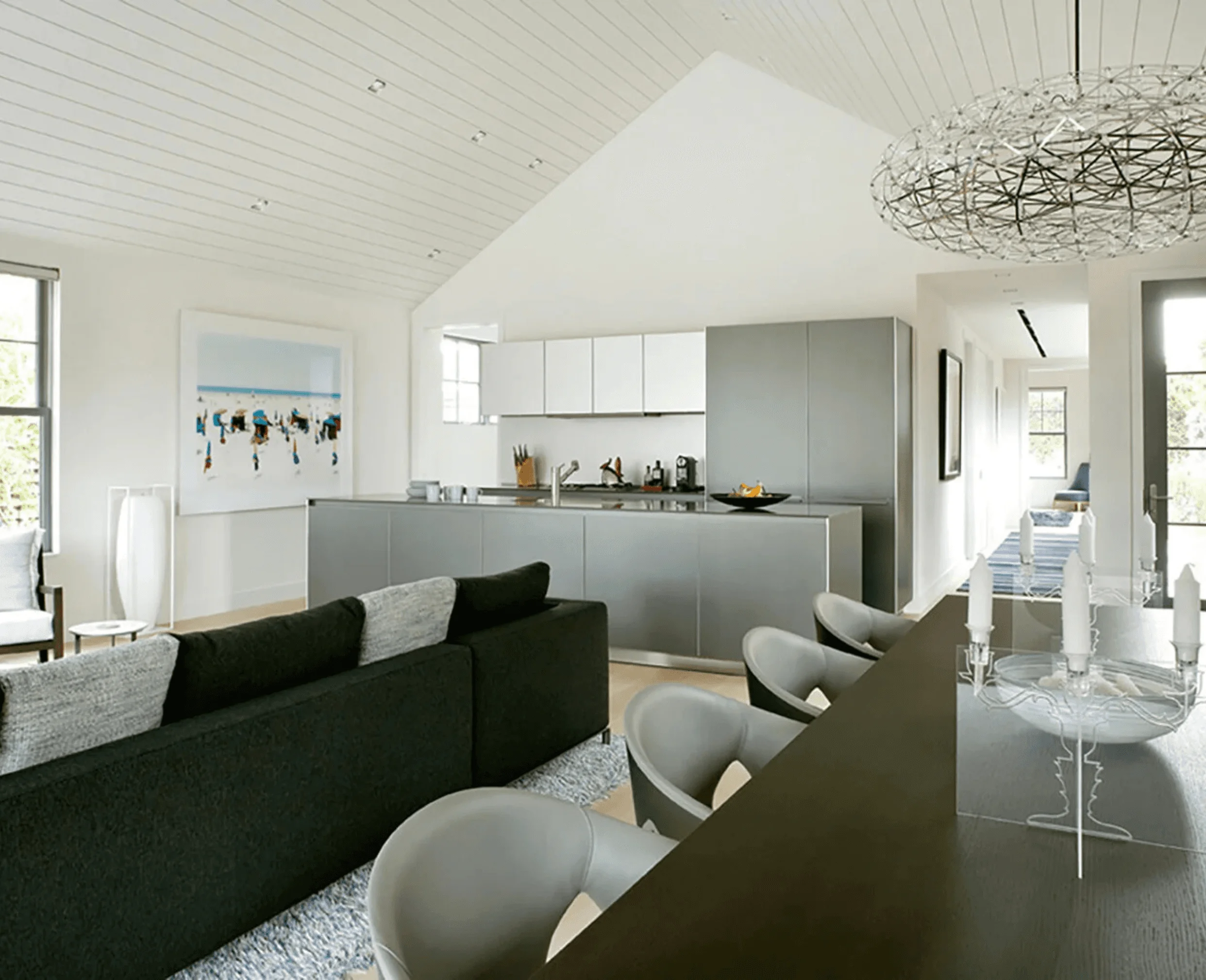 An open living and kitchen area at the Sagaponack Cottage, with a gray sectional, round dining table, and a beach-scene painting