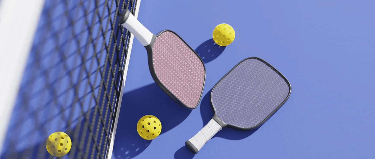 Two pickleball paddles and three yellow perforated balls are on a blue court near a net, with shadows visible on the surface.