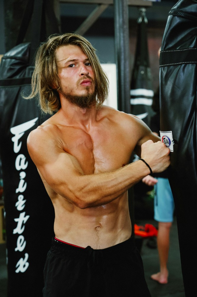 A long-haired man resting his arm on gym equipment while holding a water bottle. Quote: “I’ve tried gyms, apps, and diets – nothing compares to this program…” – Account Executive.