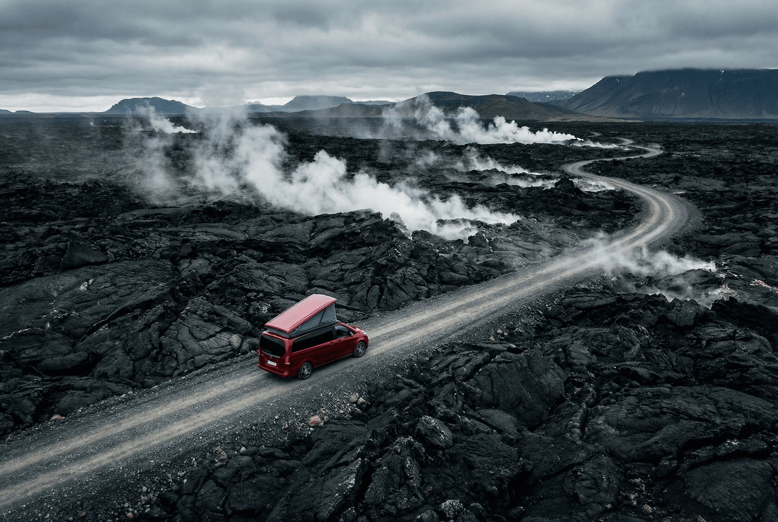 High-angle view of a red camper van driving along a winding road through a black lava field with steam vents.