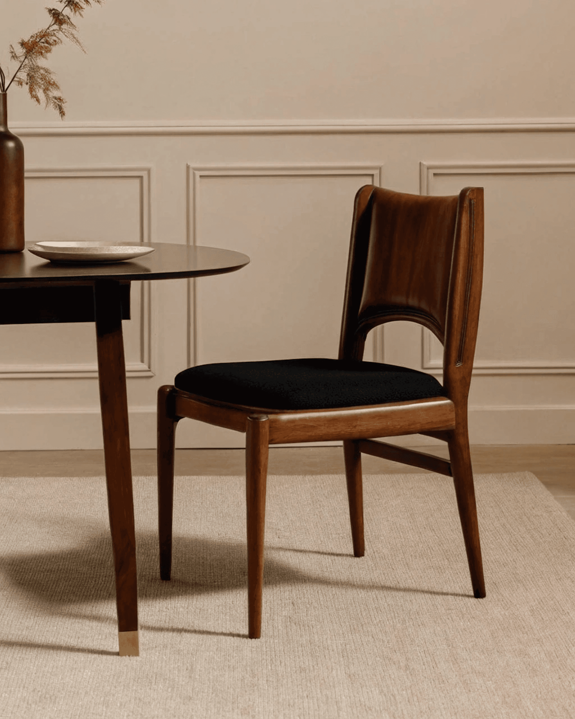 Elegant mid-century modern wooden chair with black upholstery beside a sleek black table, set against a beige paneled wall.