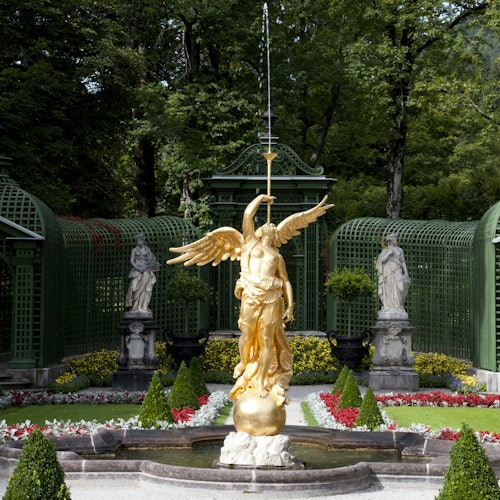 Zlatá socha anděla držícího kopí stojí v upravené zahradě, obklopená zelení a dekorativními sochami.