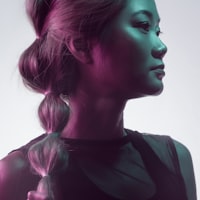 A profile portrait of a person with long hair in a ponytail, illuminated by dramatic magenta and teal lighting against a light background. They wear a dark sleeveless top and look toward the right with a contemplative expression.