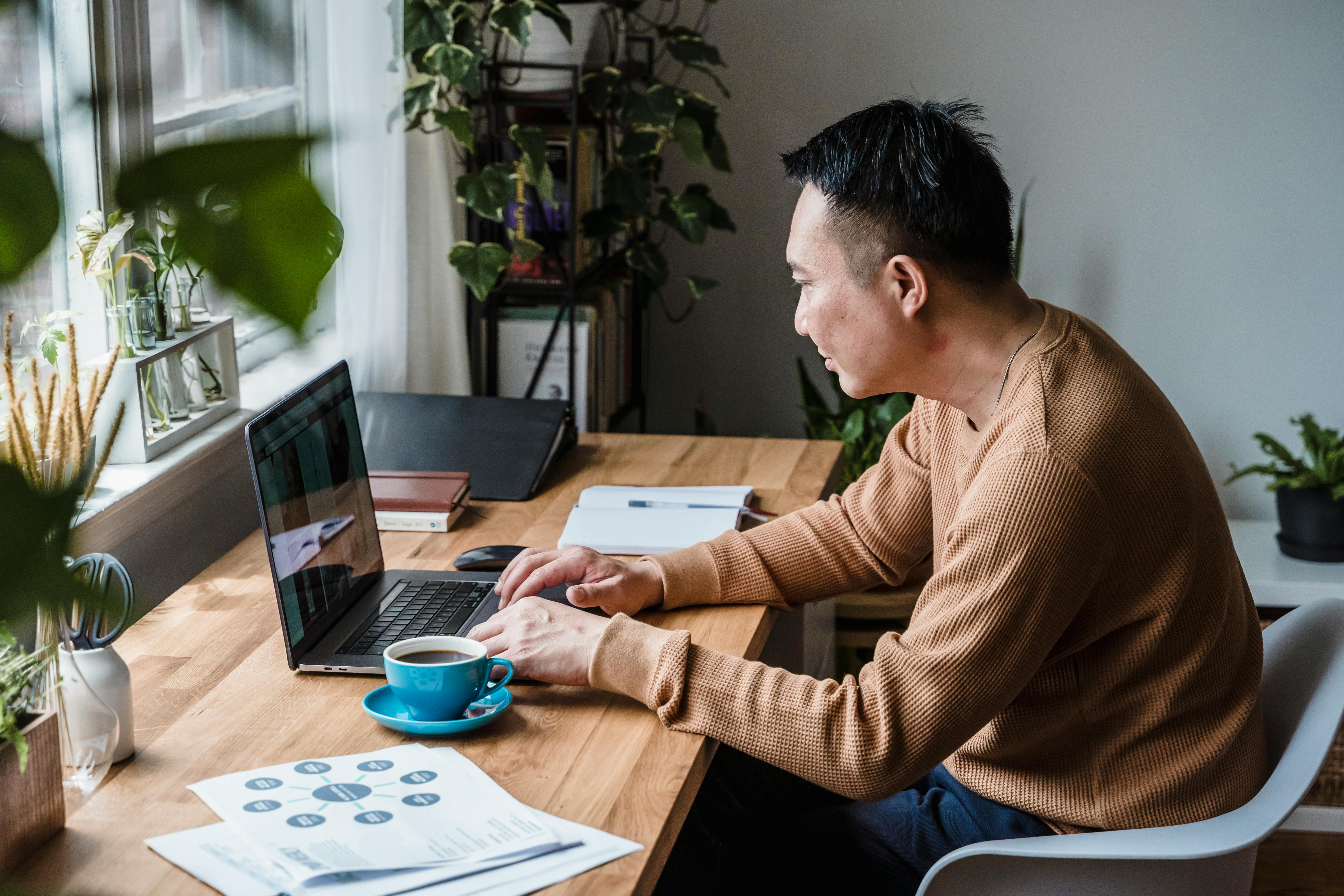 Man sitting at a desk working on a lpatop with plants, coffee and a notebook around