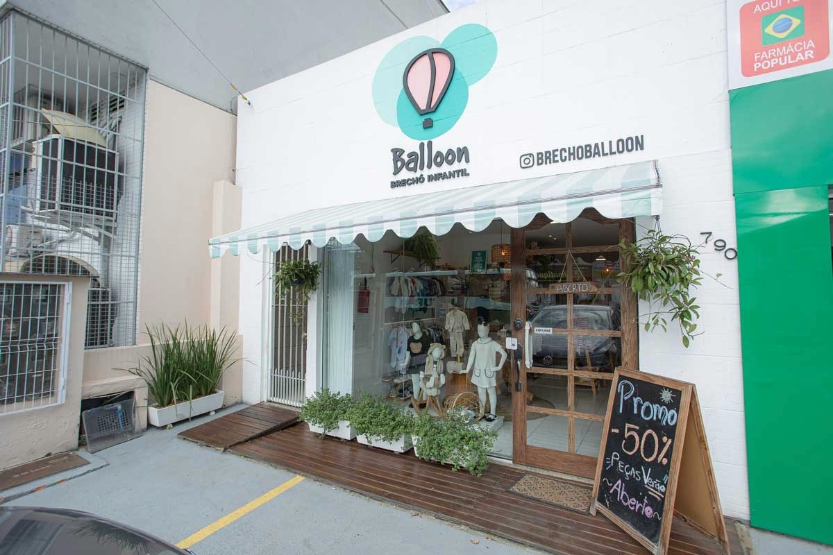 Fachada do brechó infantil, com vitrine exibindo roupas e manequins infantis, toldo listrado verde e branco, plantas decorativas e placa de promoção de 50% na entrada.