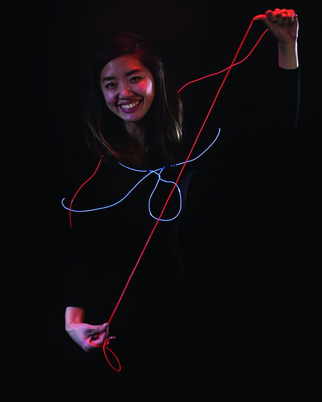 A young woman with long dark hair holds a red and blue neon string in her right hand and raises her left arm. The string forms an abstract shape resembling a bow or knot. She wears a black long-sleeved shirt and smiles at the camera. The background is entirely black.