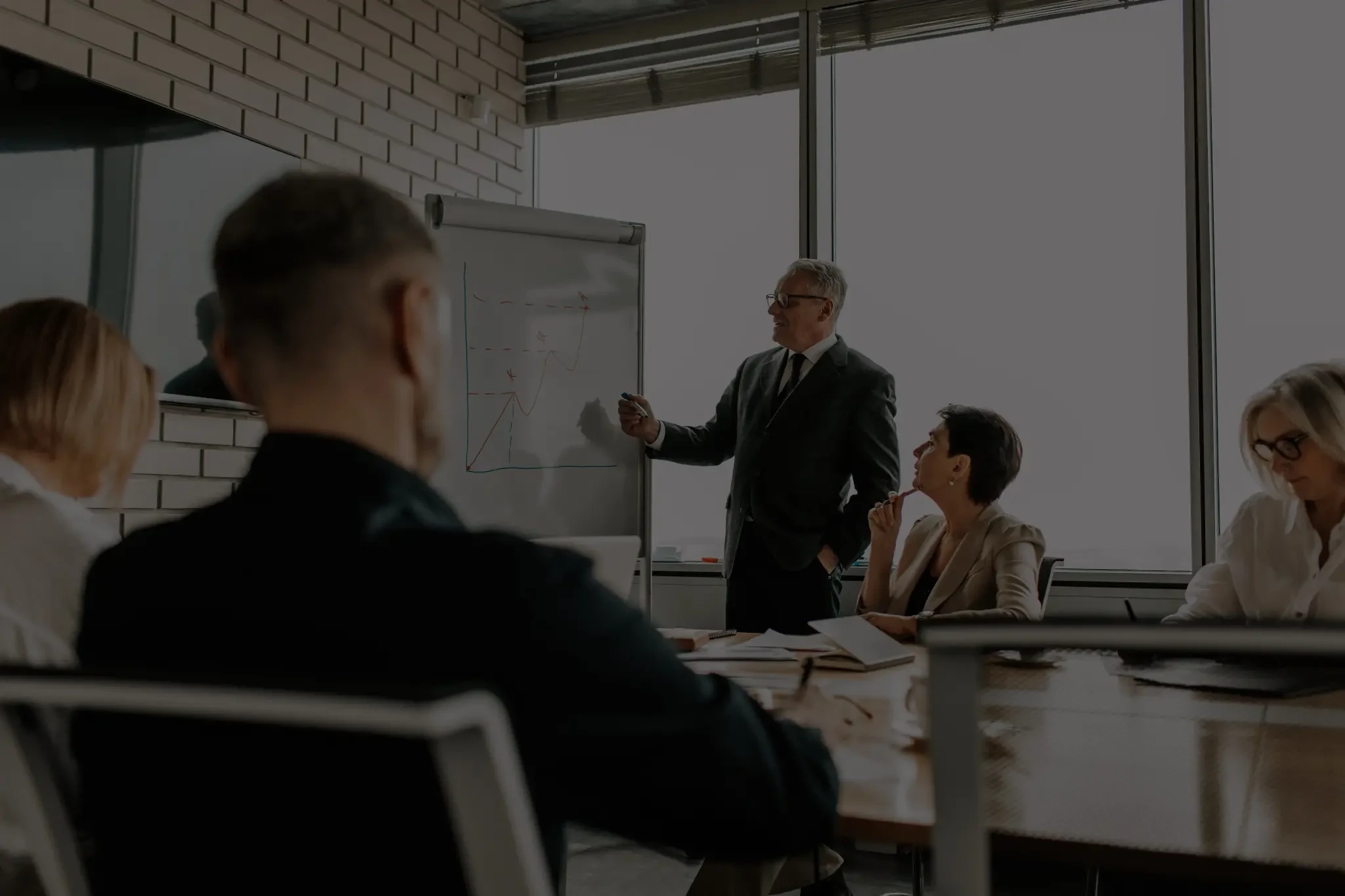 Business team listening to a presenter explain a chart on a whiteboard in a meeting room.