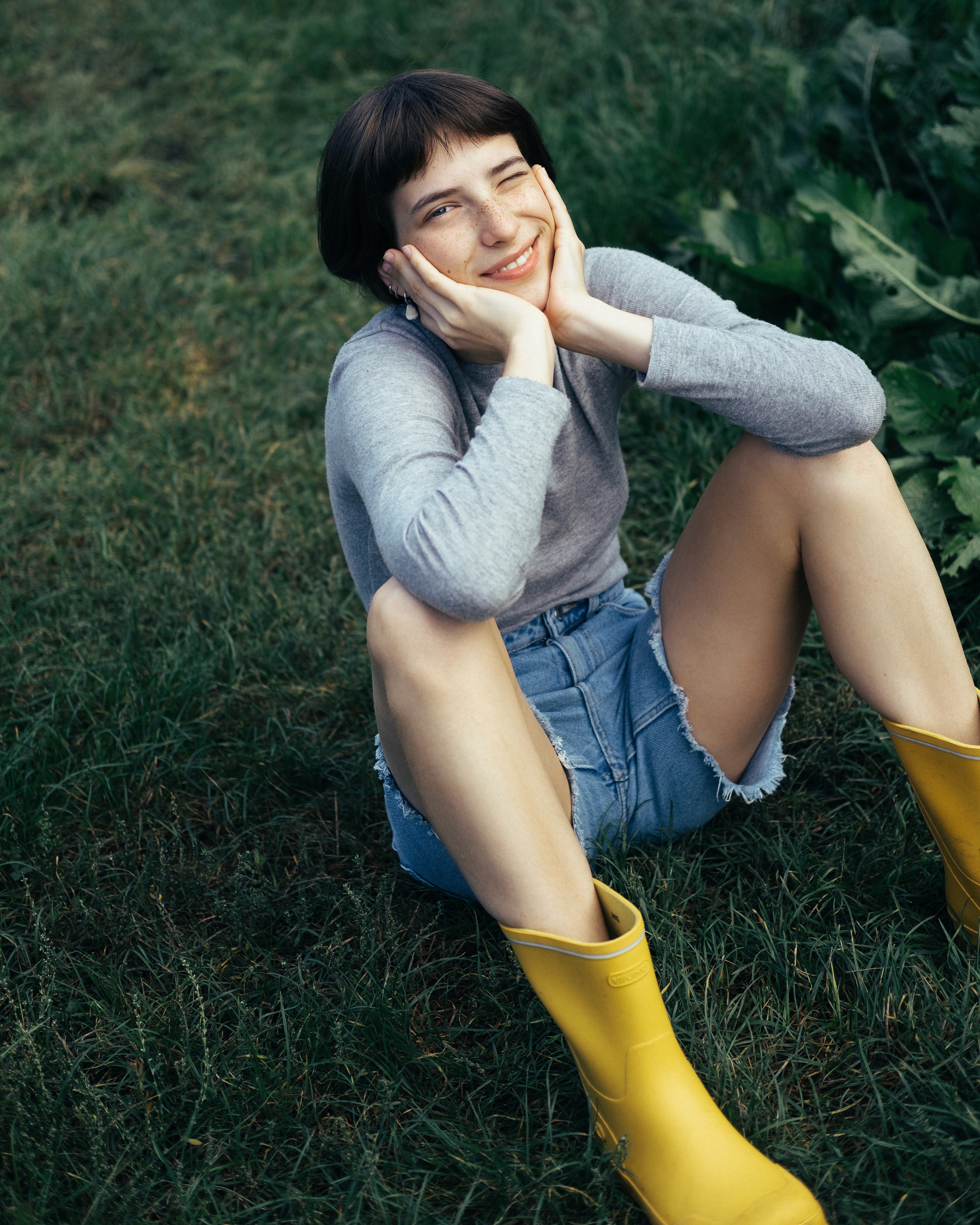 A person sitting on grass in yellow rain boots, smiling with their face resting in their hands.