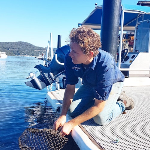 一个人在码头上跪着，手持网兜在水面上挥动。背景中可见一艘船、群山和蓝天。