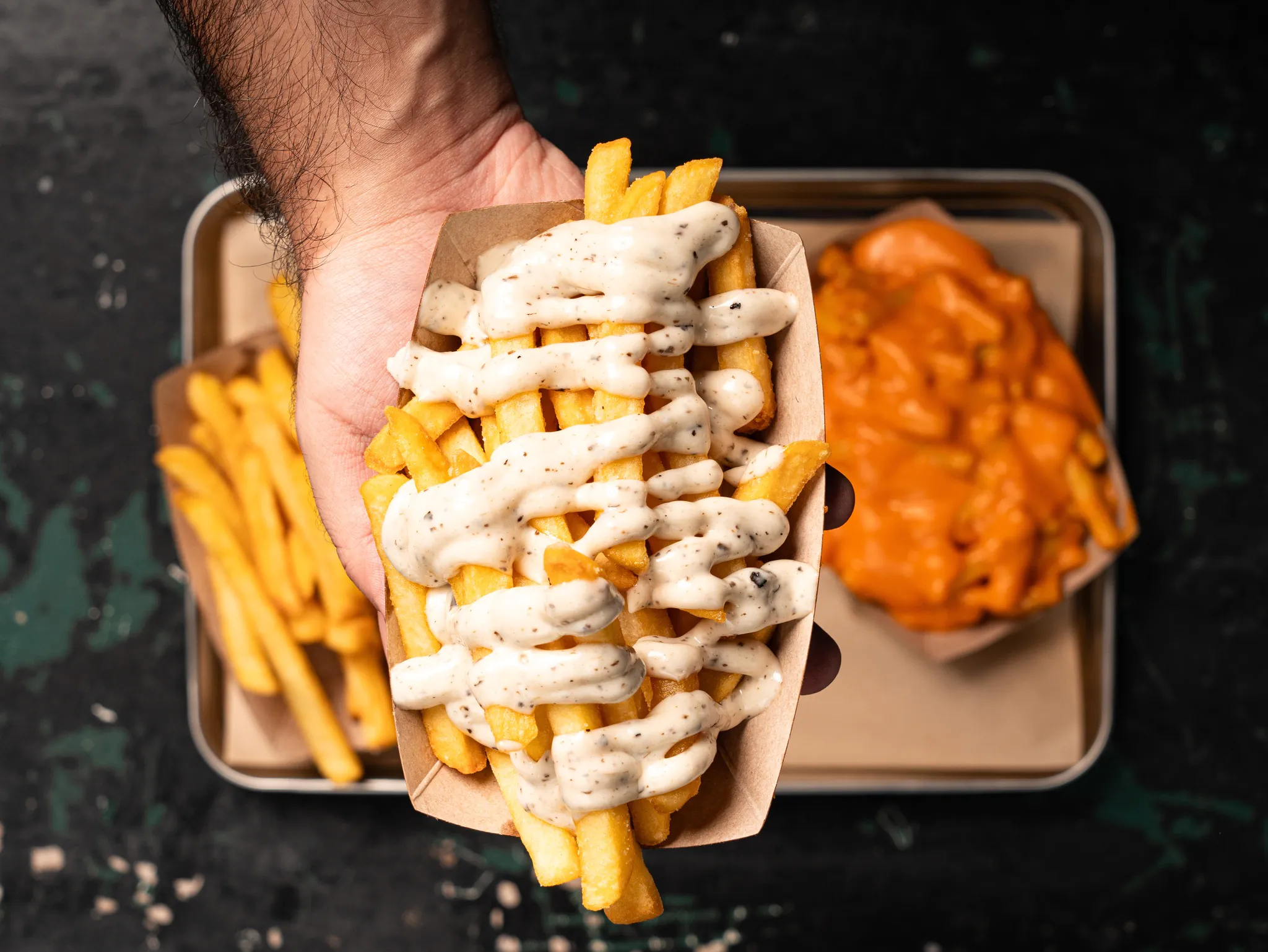 A hand holding a tray with loaded fries and sweet potato fries on a dark surface.