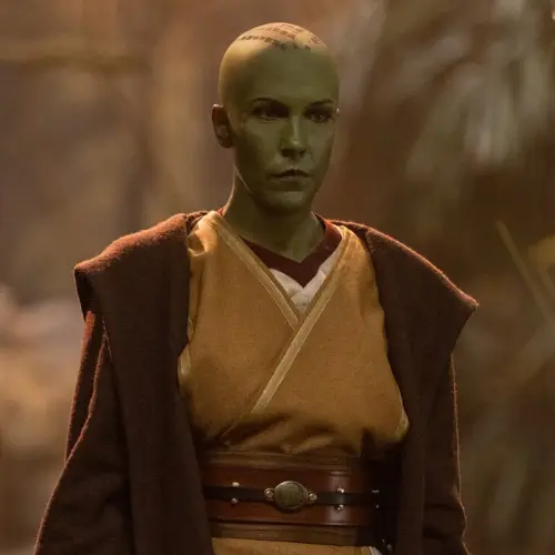 Vernestra Rwoh in a gold Jedi tabbard with a brown robe standing in front of a blurred background