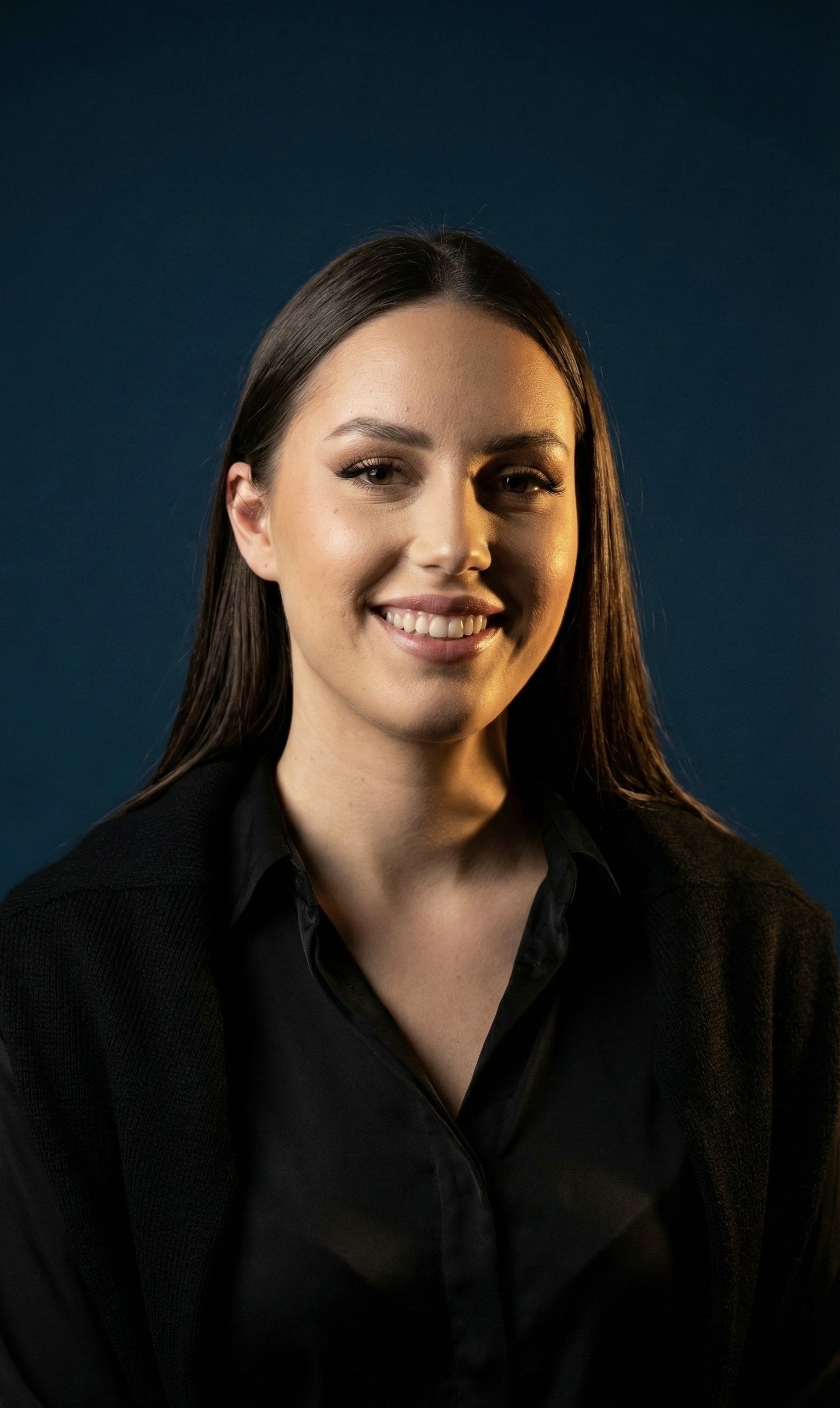 A smiling person with curly hair sits at a desk, looking at a laptop against a dark background.