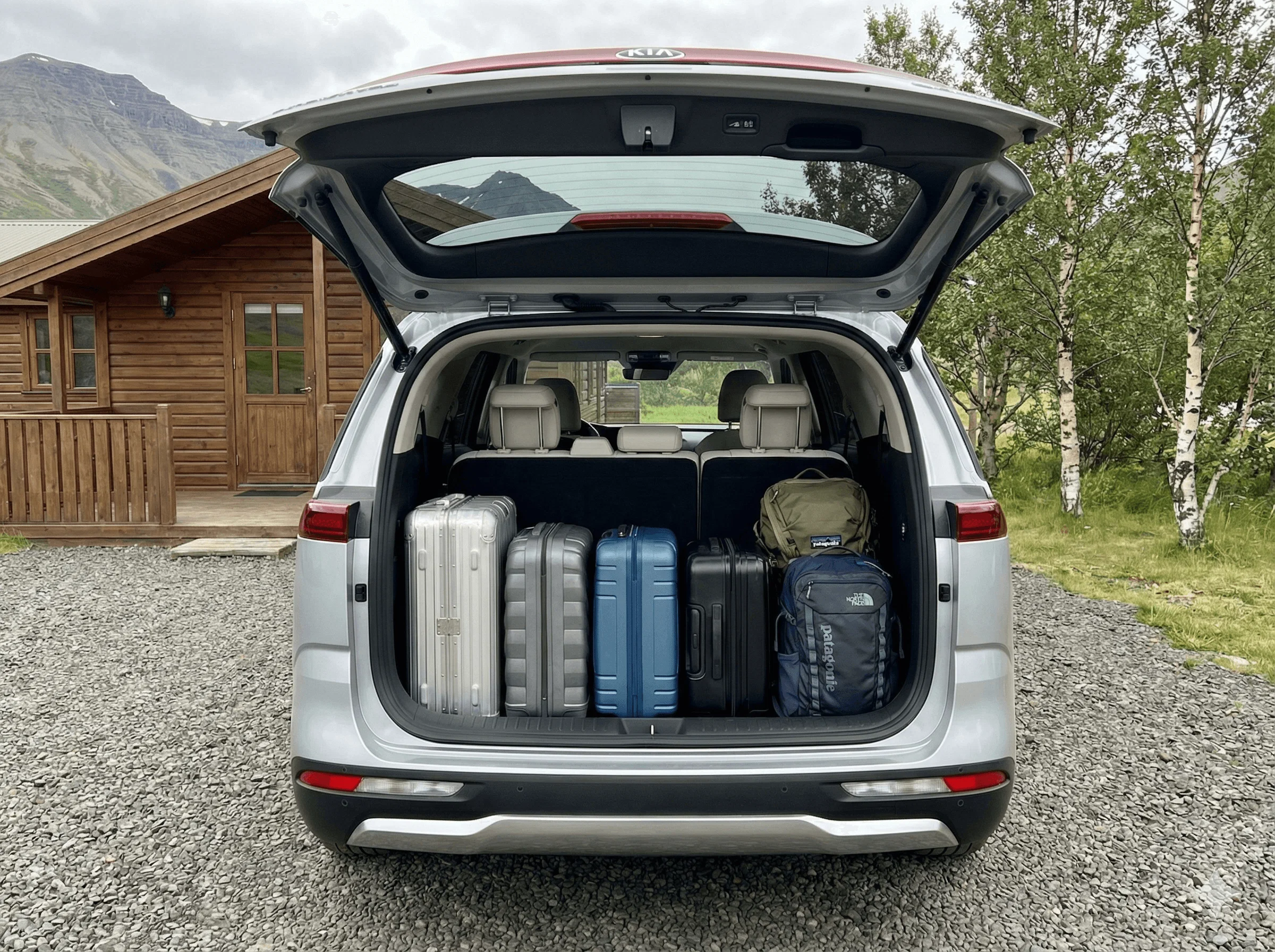 Rear view of a silver Kia SUV with the trunk open, showing it packed with suitcases next to a wooden cabin