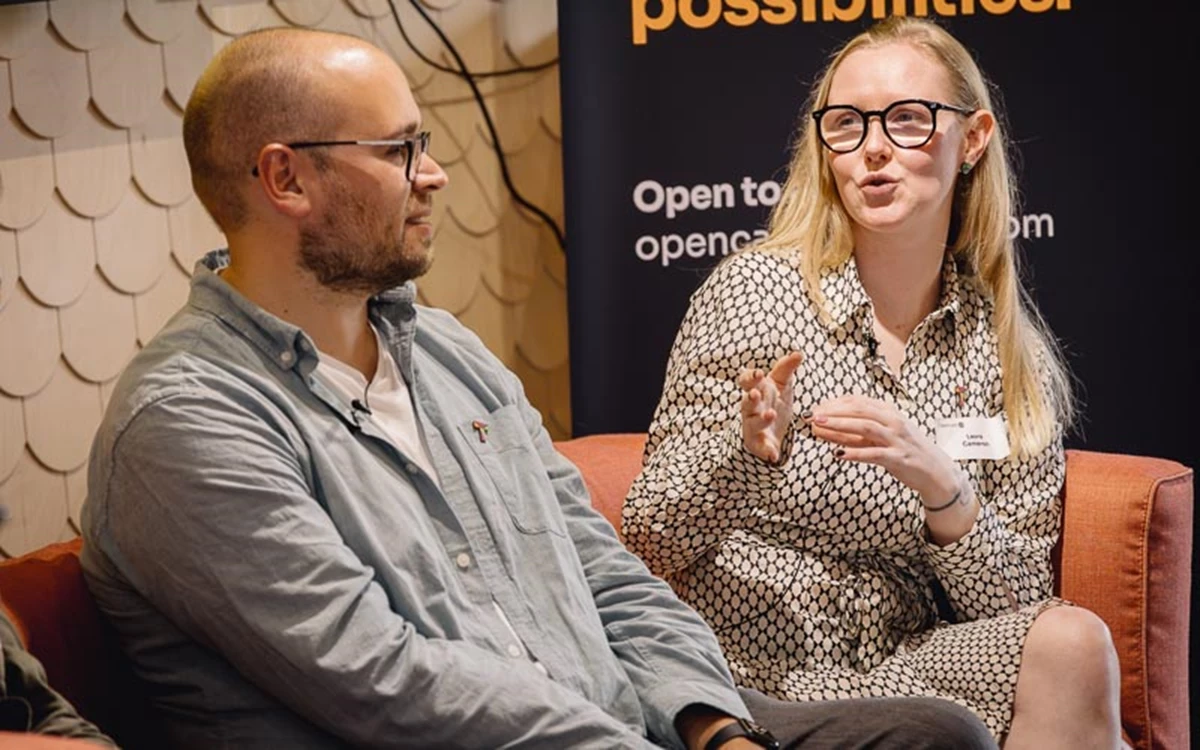 Two panelists converse on stage, with one gesturing while making a point. A branded backdrop links the discussion to a professional technology event.