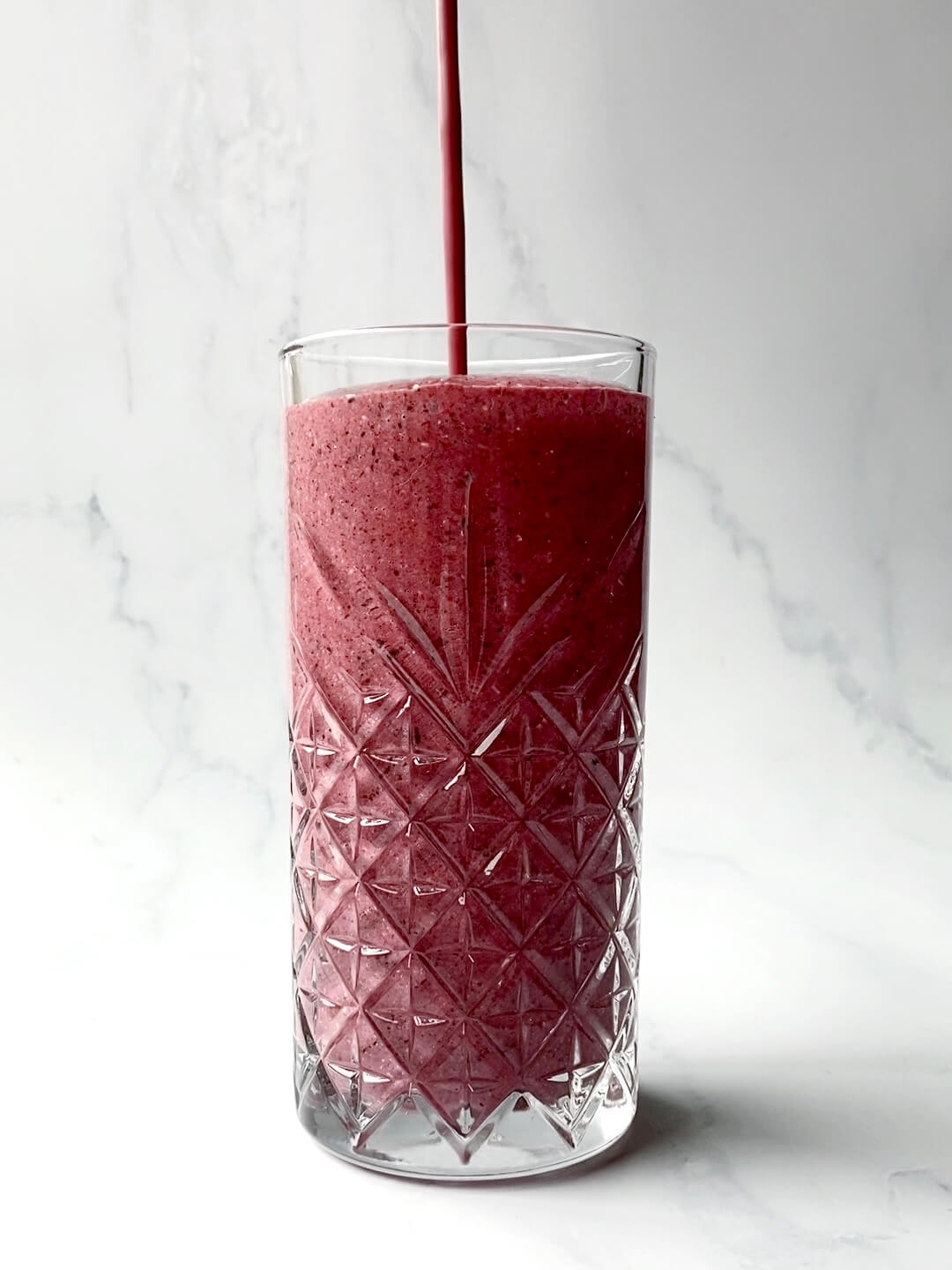 Tall glass containing a pinkish-red mixed berry smoothie that is being slowly poured from above. The glass is against a white marble backdrop