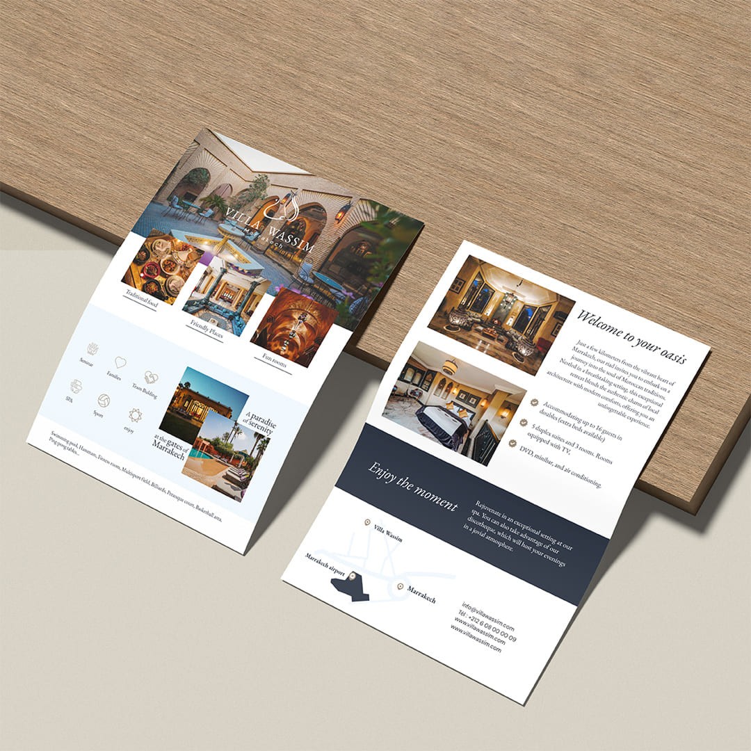 Mise en situation (mockup) de flyers imprimés conçus pour le branding de Villa Wassim Marrakech. Les deux flyers sont posés en angle sur une surface en bois. Le recto présente une composition d'images de l'hôtel (piscine, gastronomie, ambiance) et des icônes minimalistes pour les services (Spa, Sport, Team Building), avec le logo Villa Wassim en évidence. Le verso contient un texte de présentation ('Welcome to your oasis'), des photos des chambres intérieures, et les informations de contact. Ce support illustre l'expertise de Nux-Studio en design graphique de support imprimé, direction artistique et création de matériel marketing cohérent avec le branding digital du secteur hôtelier de luxe.