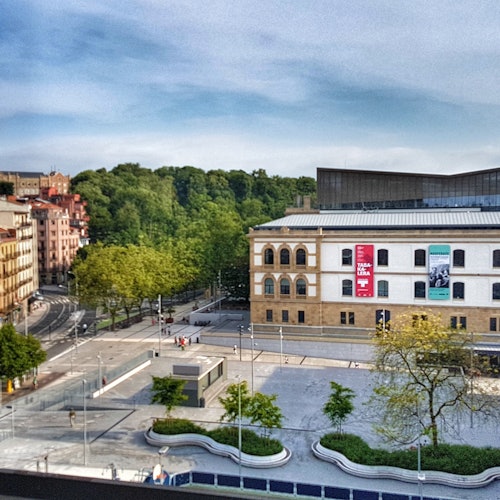 Urban scene with a plaza, modern and historic buildings, trees, and minimal people. A large building exhibits banners.
