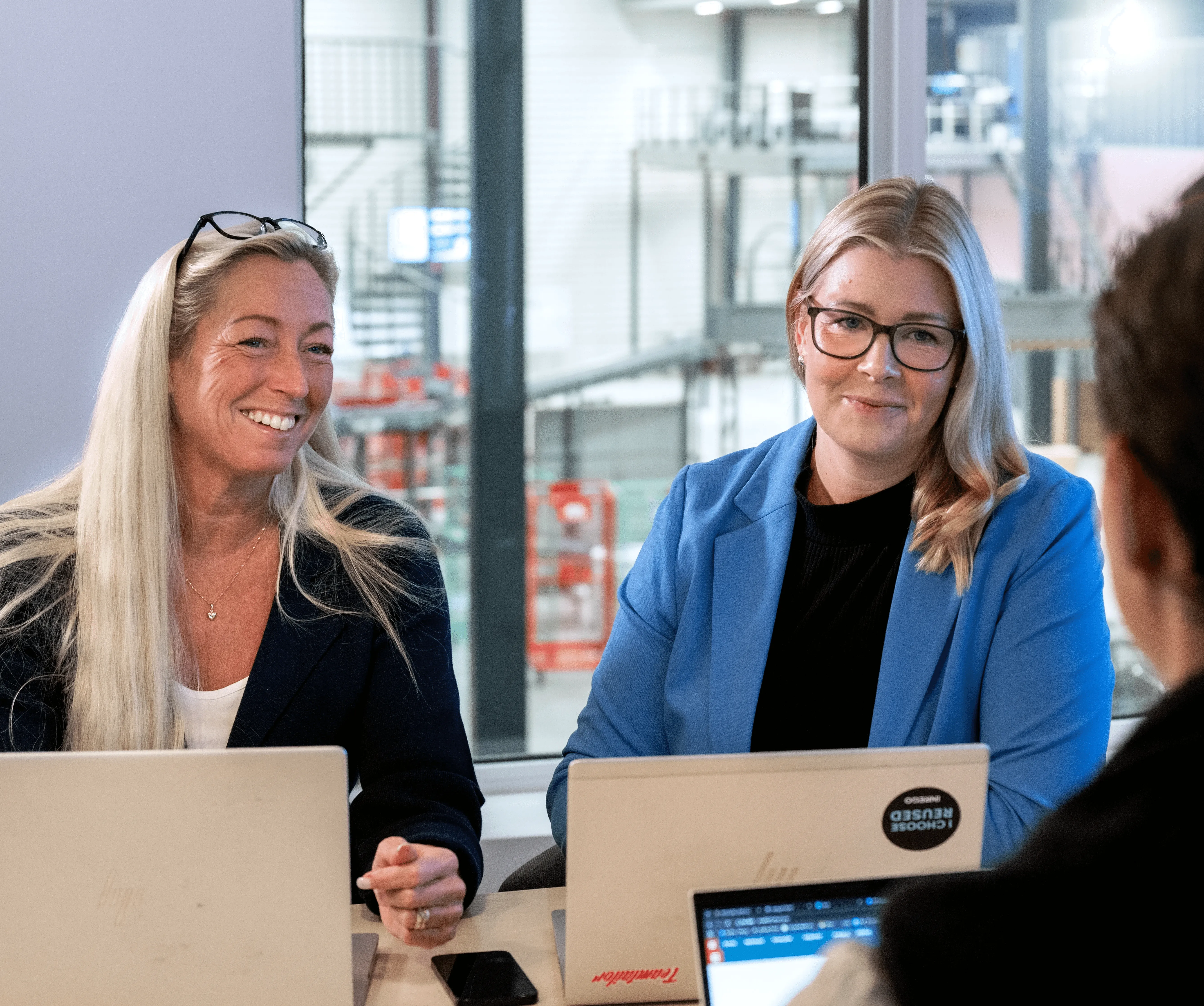 Två kvinnor sitter vid ett bord med bärbara datorer, ler och tittar mot kameran. En kvinna har glasögon och blå kavaj.