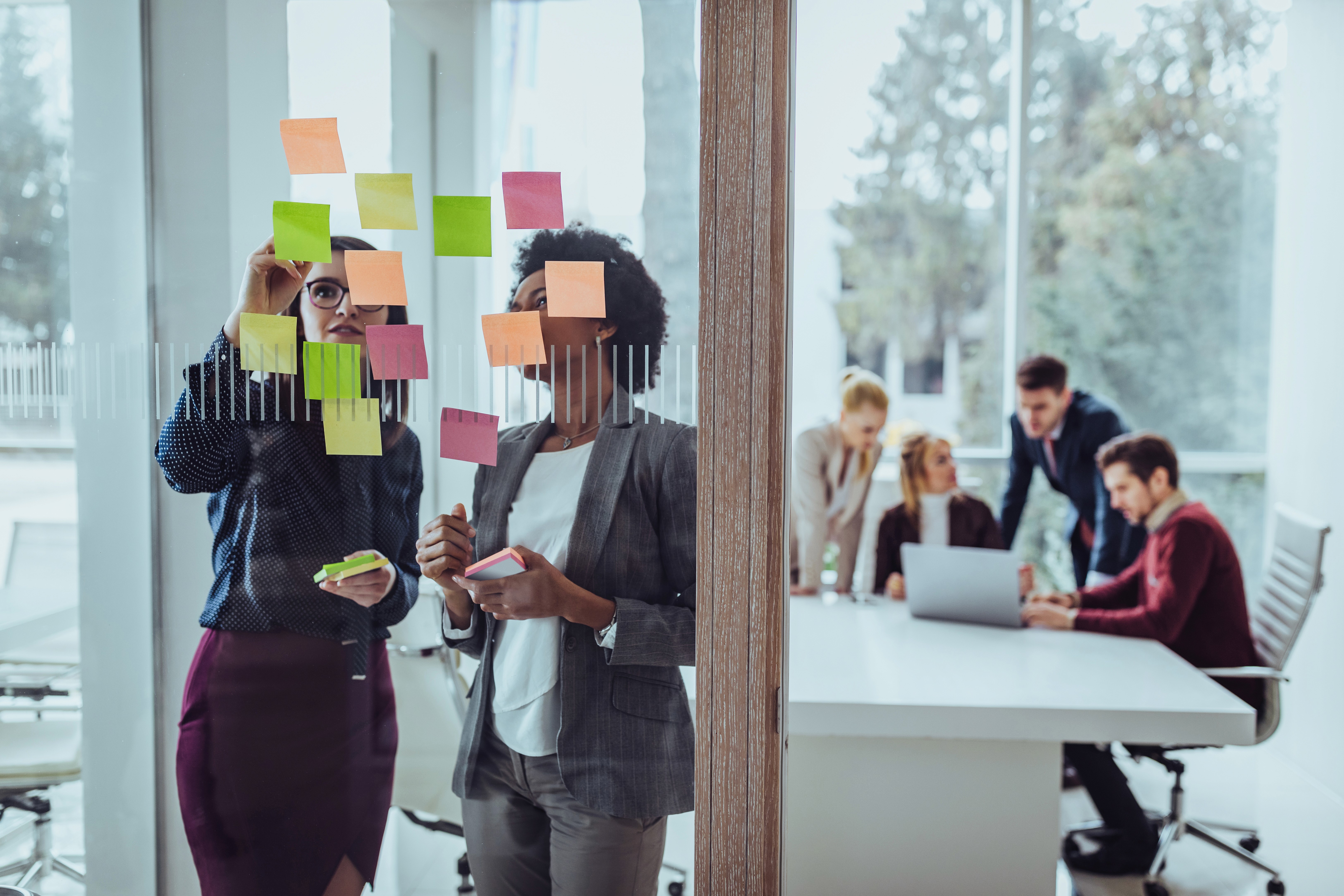 Zwei Kolleginnen brainstormen zusammen im Büro. Vor ihnen hängen mehrere Post-Its. Im Hintergrund sitzt das Team und tauscht sich miteinander aus.