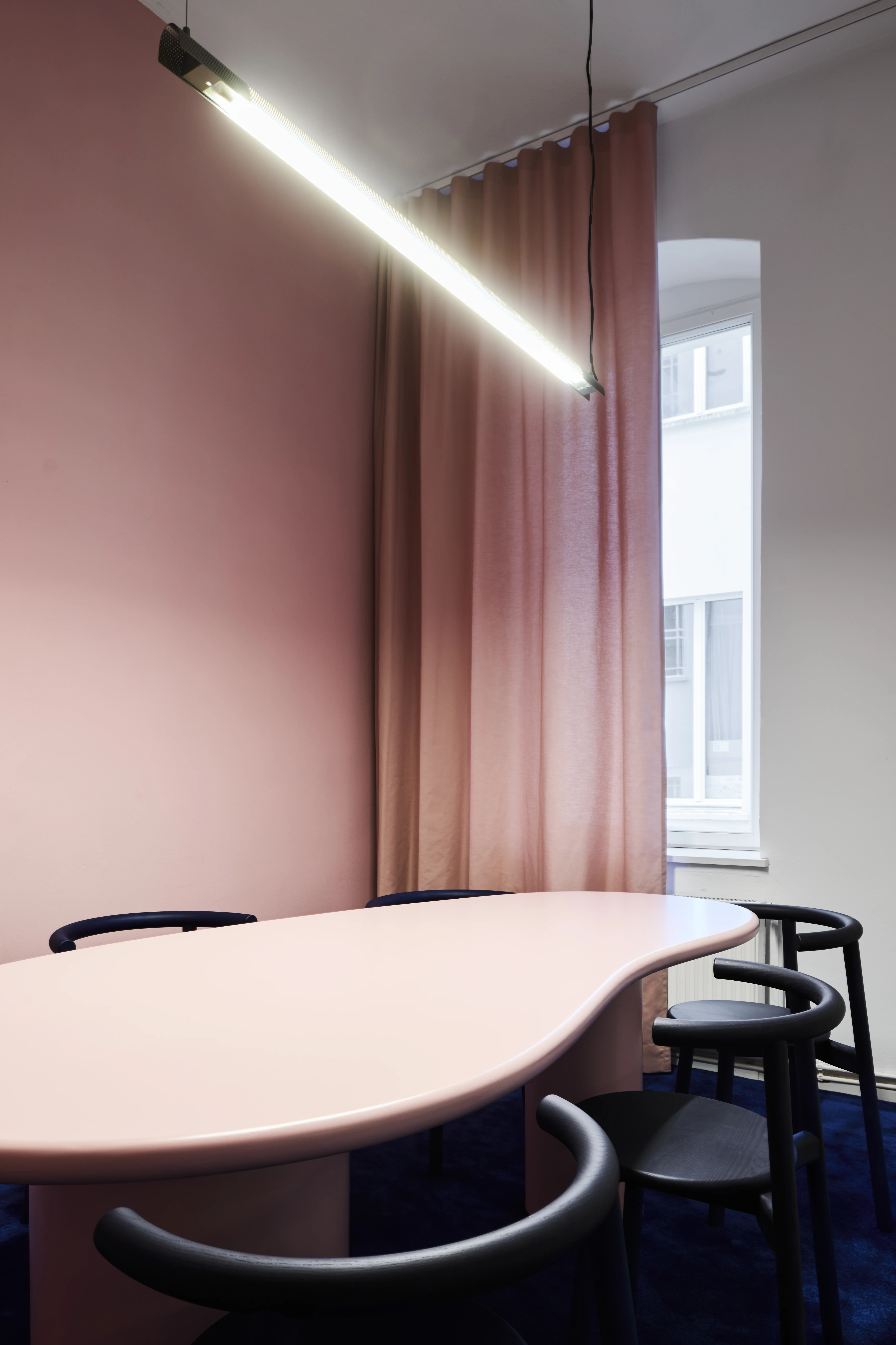 Pink conference room with organically shaped table, black chairs, tube light and rose curtain.