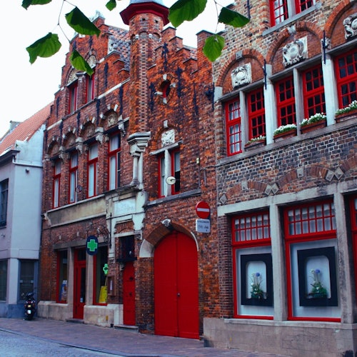 Niz starih zidanih zgrada s crvenim vratima i prozorima, od kojih jedna ima natpis ljekarne i zeleni križ. Iznand njih visi lišće. Mirna ulica.