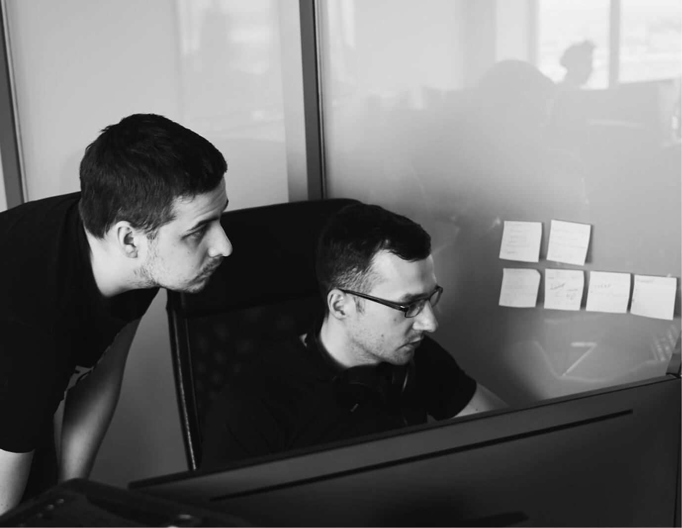 Black and white photo of two people looking at a computer screen with papers on wall